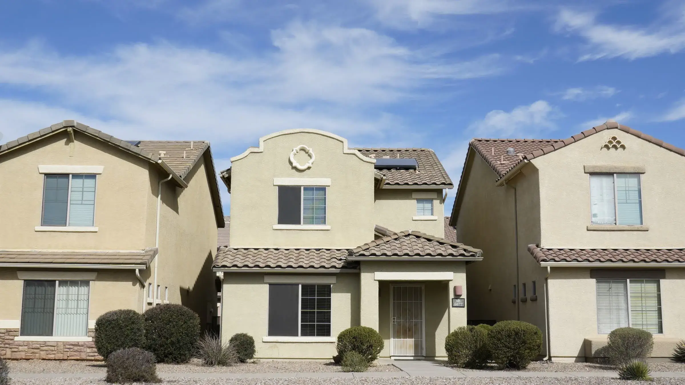 Three stucco tract houses with clay tile roofs and gravel front yards under a bright sky.