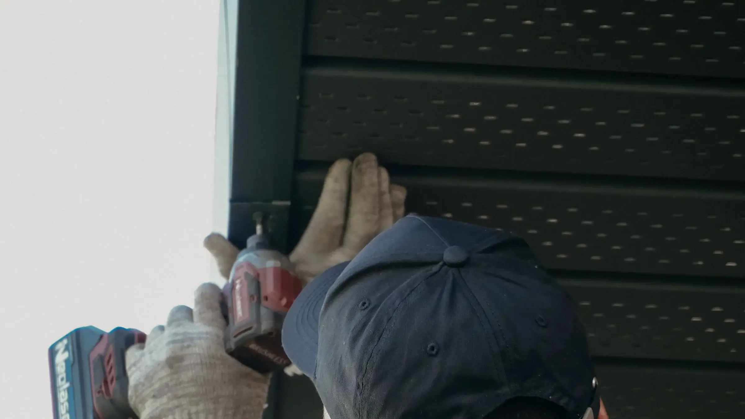 A worker in a cap and gloves driving a screw overhead with a cordless drill.